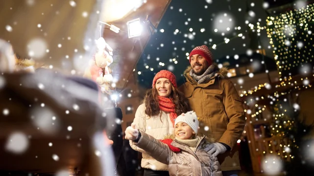 family, winter holidays and celebration concept - happy mother, father and little daughter at christmas market on town hall square in tallinn, estonia over snow